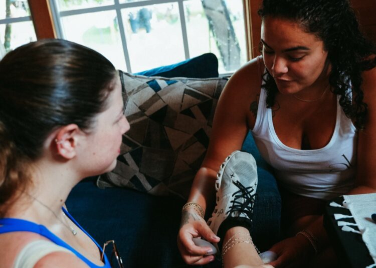 Two women sitting indoors, one applying a bandage to the other's arm, with a window showing outdoor greenery behind them.