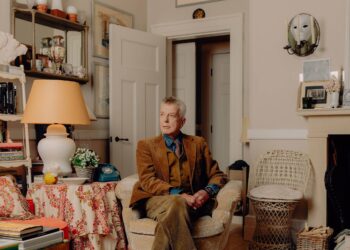 Nicky Haslam sentado en el salón de su casa en Cotswolds con chaqueta y pantalones marrones