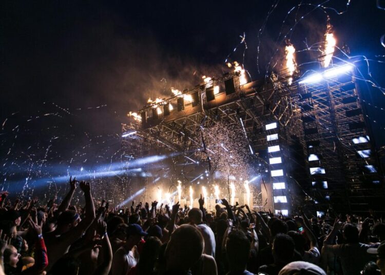 A massive outdoor festival crowd at night with arms raised toward a large stage structure topped with flames and confetti cannons firing silver streamers into the air, with bright white and amber stage lights cutting through the smoke and darkness above the packed crowd.