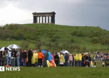 A long queue of people in lots of different coloured raincoats and holding several umbrellas. They are standing in a field with hills in the background. At the top of the hill is the Penshaw Monument which was built in the style of an ancient Greek temple.