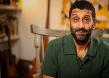 Adeel Akhtar estaba sentado en una silla, sonriendo al frente, mientras viste una camisa verde oscuro. Tiene cabello negro y barba con colores grises.