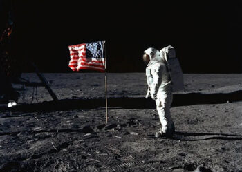 Un astronauta con traje espacial se encuentra en la Luna junto a una bandera estadounidense, con partes del módulo de aterrizaje lunar y huellas visibles en la superficie gris y polvorienta contra un cielo negro.