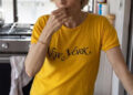 una mujer vestida de amarillo "Nueva York" camiseta parada en una cocina, sosteniendo un trozo de melón en la boca. En el mostrador, frente a ella, hay trozos de melón cortados y un cuchillo.