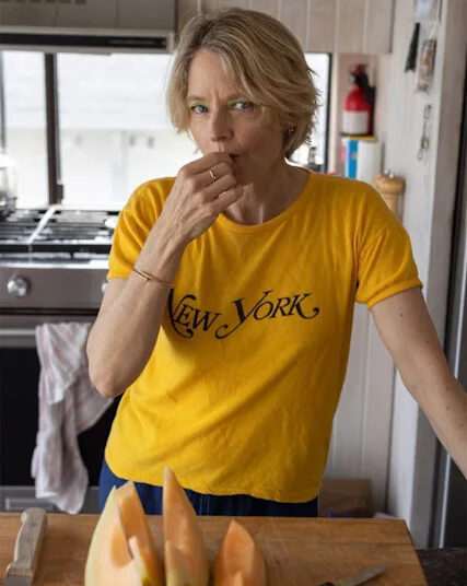 una mujer vestida de amarillo "Nueva York" camiseta parada en una cocina, sosteniendo un trozo de melón en la boca. En el mostrador, frente a ella, hay trozos de melón cortados y un cuchillo.