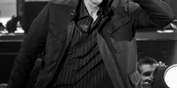 Un hombre con camisa y chaqueta a rayas se inclina hacia adelante en el escenario, se lleva una mano a la oreja y hace una expresión facial animada y juguetona. La imagen está en blanco y negro.
