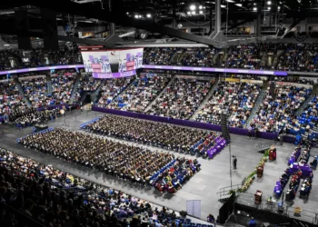Vista amplia de una ceremonia de graduación en un estadio cubierto abarrotado, con filas de graduados sentados en el suelo y un escenario instalado al frente.