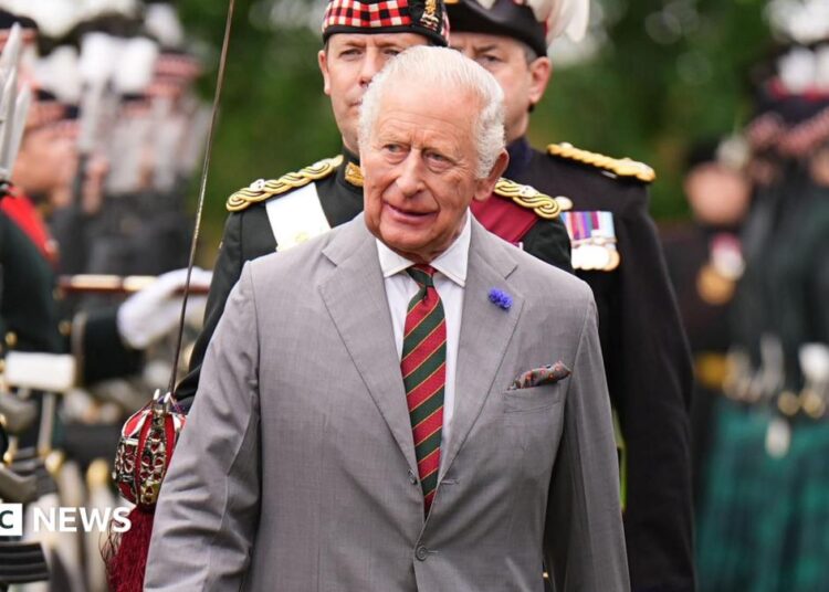 King Charles III wearing a grey suit with a white shirt and red and green striped tie. He is walking through the Palace Guard - a line of soldiers, wearing black uniforms.