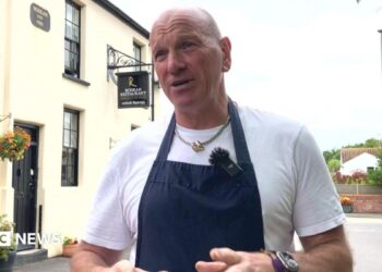 A restaurant owner wearing a navy blue apron and white t-shirt stands outside his restaurant while explaining the impact traffic will have on his business