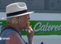 A woman with blonde hair standing next to an ice cream van. She is wearing a straw hat and a denim top. She is turned to the side facing towards the right and is eating an ice cream in a cone.