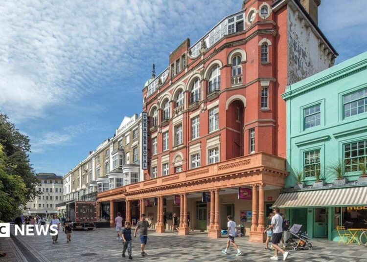 A large red-brick theatre building with cream detailing and arched windows stands on a pedestrian street, with people walking past and colourful shop fronts beside it under a bright sky.