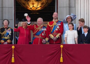 La familia real británica vestida de manera oficial se encuentra en un balcón saludando a la multitud.