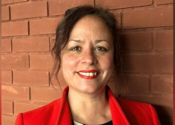 Michelle Daurat tiene cabello y ojos castaños y su cabello está recogido. Lleva un lápiz labial rojo, que combina con su abrigo, y un top negro. Ella está sonriendo.
