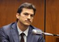 Un hombre con traje, corbata y bigote está sentado en una sala del tribunal, luciendo serio, con un gran micrófono frente a él y paredes con paneles de madera al fondo.