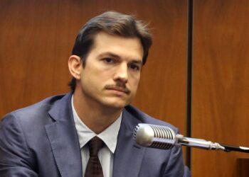Un hombre con traje, corbata y bigote está sentado en una sala del tribunal, luciendo serio, con un gran micrófono frente a él y paredes con paneles de madera al fondo.