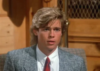 A young man with blond hair wearing a gray suit jacket, white shirt, and red tie sits indoors, looking slightly surprised. Wood paneling and a mesh-covered cabinet are in the background.