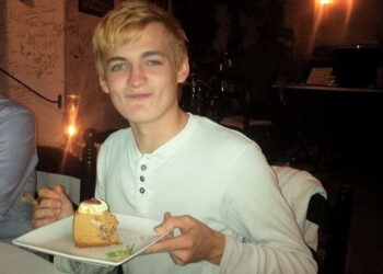 Un joven de cabello rubio, vestido con camisa blanca, sonríe mientras sostiene un plato con un trozo de pastel y un tenedor, sentado en un restaurante con poca luz.