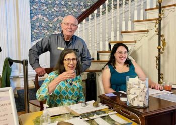 A smiling woman holds a drink at a table with papers and a clear container, while another woman sits beside her, both at a registration or information desk with a man standing behind them in a hallway with decorative wallpaper.