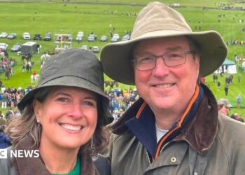 Alice Dearing and her husband John stood next to each other having their picture taken outside. They are dressed in coats and hats each and are looking directly at the camera and smiling. Alice is on the left and John is on the right. They are at an event with crowds of people in the background.