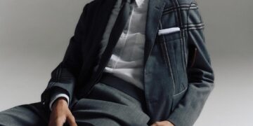 Un joven con un elegante traje gris sentado en una silla de madera sobre un fondo neutro.