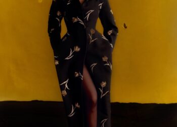 Model in a floral black coat with a thigh-high slit and red heels, surrounded by butterflies against a yellow background.