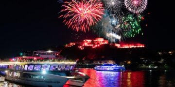 Un barco flota en un río en primer plano mientras los fuegos artificiales explotan sobre un castillo iluminado al fondo por la noche.