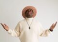 Nick Cooper lleva un suéter de cuello alto color crema y un sombrero de ala ancha a juego mientras mira hacia arriba con ambos brazos levantados durante una sesión de retratos sobre un fondo blanco.