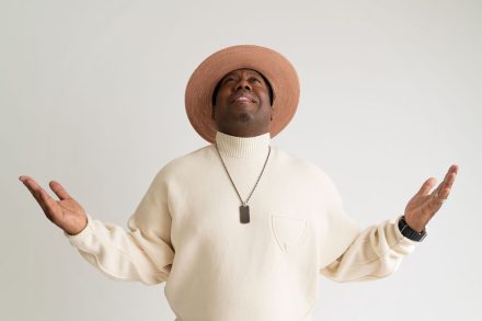 Nick Cooper lleva un suéter de cuello alto color crema y un sombrero de ala ancha a juego mientras mira hacia arriba con ambos brazos levantados durante una sesión de retratos sobre un fondo blanco.