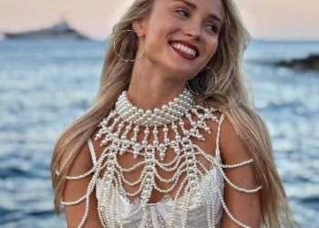 Mujer de cabello rubio con un collar de perlas blancas y un top blanco junto al océano.