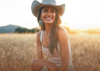 La cantante de country Annie Bosko, con sombrero de vaquero, está sentada en un campo iluminado por el sol, sonriendo con las montañas de fondo.
