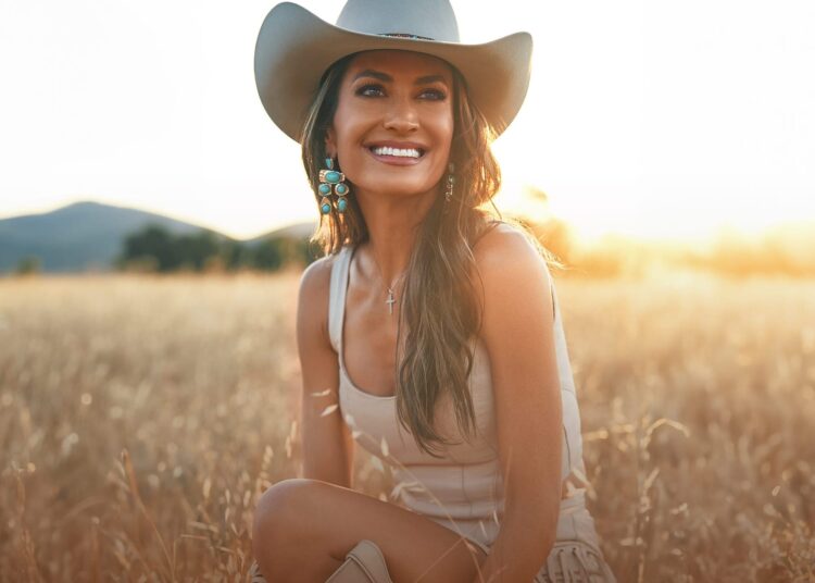 La cantante de country Annie Bosko, con sombrero de vaquero, está sentada en un campo iluminado por el sol, sonriendo con las montañas de fondo.